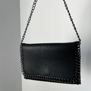 Svart väska  - Väskan kan användas både som crossbody eller clutch då kedjan smidigt går att stoppa in i väskan.  Insidan har ett fack med dragkedja samt en utan.