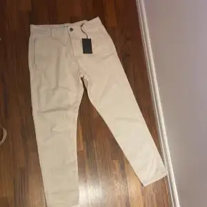 Säljer ett par snygga beige chinos från Only & Sons. De är i storlek W28 L32 och är helt nya med taggar kvar. Perfekta för både vardag och lite mer uppklädda tillfällen. Materialet är mjukt och bekvämt, och de har en klassisk passform med raka ben. Färgen är ecru, vilket är en ljus beige nyans. Passa på att fynda dessa stilrena byxor!