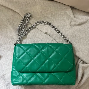 Grön quiltad axelväska med kedja - Säljer en snygg grön axelväska i quiltat mönster. Väskan har en silverfärgad kedja som axelrem och är perfekt för både vardag och fest. Den är i bra skick och redo för en ny ägare!