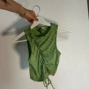 Grön ribbad linnetopp från Divided - Säljer en snygg grön ribbad linnetopp från Divided. Toppen är ärmlös och har en cool snörningsdetalj på framsidan. Perfekt för sommaren och kan stylas både till vardags och fest. Toppen är i nyskick och redo för en ny ägare!