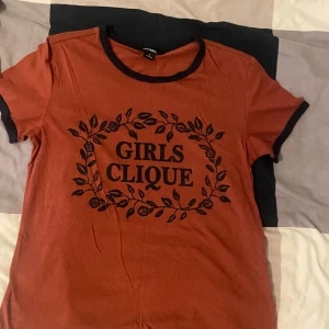 Röd t-shirt från Monki med texten 'Girls Clique' - Säljer en snygg röd t-shirt från Monki med texten 'Girls Clique' i svart. T-shirten har korta ärmar och en rund halsringning med svarta detaljer. Perfekt för vardagsbruk eller en avslappnad dag ute. Den är i bra skick och väldigt bekväm att ha på sig. Passar perfekt till jeans eller shorts!