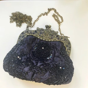 Svart kuvertväska med pärlor och paljetter - Säljer en superfin svart kuvertväska med vackra pärlor och paljetter. Väskan har en antik guldig kedja och detaljer på ramen som ger den en vintage look. Perfekt för fest eller bal! Väskan är i nyskick och har en praktisk stängning med en rhinestones på toppen. Insidan är fodrad med svart tyg och har  litet fack för förvaring.