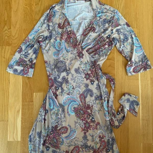 Beige blommig omlottklänning i strl L från Revd’elle - Sparsamt använd och i fint begagnat skick.  Tyvärr har jag klippt bort lappen med storlek och material men tror det är en L, dubbelkolla måtten nedan.  Mått Över bröstet: ca 50 cm Längd från axeln och ner: ca 96 cm 