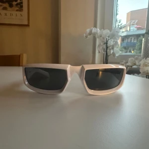Vita solglasögon från Prada - Säljer ett par snygga vita solglasögon från Prada. De har en rektangulär form och svarta linser. Perfekta för att ge en cool och modern look. Passar både till vardags och fest.
