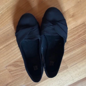 Svarta ballerinaskor med knutdetalj - Säljer ett par snygga svarta ballerinaskor med en fin knutdetalj på ovansidan. De är perfekta för både vardag och fest. Skorna är i syntetmaterial och har en bekväm passform. Storleken är inte angiven, men de ser ut att vara i bra skick.