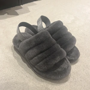 Mysiga grå sandaler med päls - Säljer ett par supermysiga grå sandaler med fluffig päls. De har en bred rem över foten och en elastisk rem runt hälen för extra komfort. Perfekta för att hålla fötterna varma och sköna hemma.