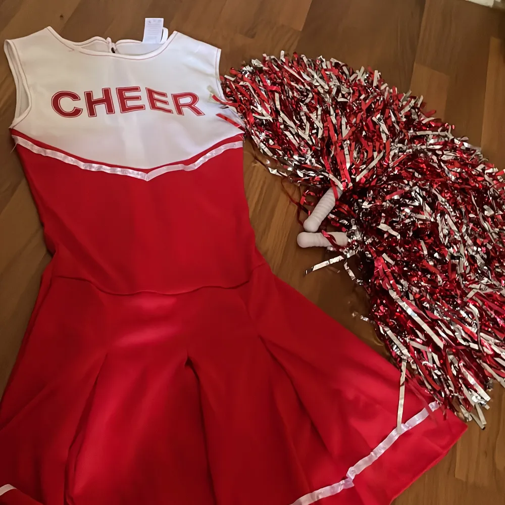 Säljer en superfin röd cheerleaderklänning med texten 'CHEER' på bröstet. Klänningen är ärmlös och har en dragkedja på baksidan. Den är i nyskick och perfekt för fest eller andra roliga aktiviteter. Pom-poms i matchande färger ingår också!. Klänningar.