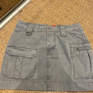 Grå kjol från Divided - Säljer en snygg grå kjol från H&M Divided. Den har en cool design med flera fickor och detaljer som gör den unik. Kjolen är i bomull och har en normal passform. Perfekt för vardagsbruk eller en avslappnad dag ute. Den är i bra skick och redo för en ny ägare!