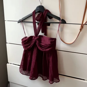 Vinröd topp med knytband - Säljer en superfin vinröd topp med knytband. Toppen är ärmlös och har en söt rosett framtill. Den är gjord av ett lätt och luftigt material, perfekt för sommarens fester. Baksidan har en smockad detalj som gör att den sitter bekvämt och snyggt.