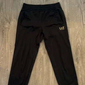 Svarta mjukisbyxor från Emporio Armani - Säljer ett par svarta mjukisbyxor från Emporio Armani. Byxorna har en elastisk midja och två fickor fram. De har en EA7-logga på vänster ben. Perfekta för en avslappnad och bekväm stil.