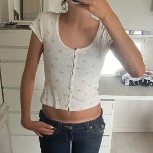 Vit kortärmad topp med knappar - Säljer en söt vit kortärmad topp med små blommor och knappar framtill från Brandy Melville. Inga defekter! Storleken är ju one size men toppen passar xs-m skulle ja säga då den är stretchig