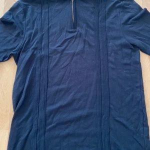 Mörkblå t-shirt från Le Breve - En mörkblå t-shirt från Le Breve med korta ärmar och dragkedja vid halsen. T-shirten har en enkel design med vertikala sömmar på framsidan.