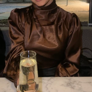 Satinblus - Jättefin brun satinblus som inte längre går att köpa. Inga skador eller skavanker.  Blusen har en hög krage med plisserade detaljer på axlarna, två knappstängningar bakom nacken med en öppen detalj, något, pösiga långa ärmar. 