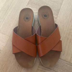 Bruna sandaler från Birkenstock - Ett par bruna sandaler från indiskan med korsade remmar i skinn. Sandalerna har en bekväm fotbädd och en vit sula med räfflad kant.
