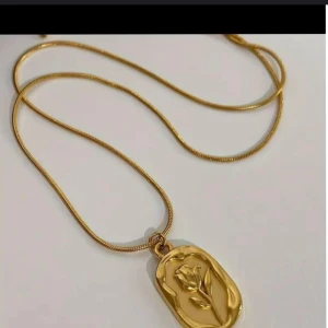 Guldhalsband med blomma - Ett elegant guldhalsband med ett hänge som föreställer en blomma. Hänget har en oregelbunden form och en relief av en blomma i mitten. Kedjan är tunn och guldpläterad.