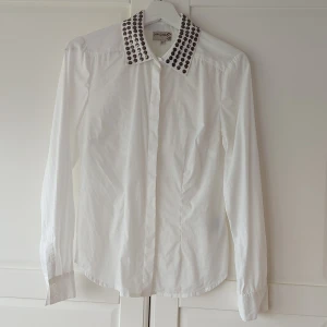 Vit skjorta från Kön & Mön - En snygg vit skjorta/blus från Kön & Mön med långa ärmar och en unik krage dekorerad med runda metallknappar. Skjortan har en broderad detalj på ärmen och knäppning framtill. Lätt insvängd och så snygg till jeans! Sparsamt använd.
