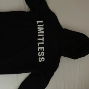  Svart 'LIMITLESS' Hoodie Med Luva - En svart hoodie med texten 'LIMITLESS' tryckt i vitt på framsidan. Hoodien har en luva med dragsko och långa ärmar. säljer på grund av storlek men passa för både L och M 