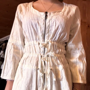Klänning - Vintage klänning med mörkgrå dragkedja i crémefärgat vit. Går ner till knäna på mig som är 166 cm lång. Storlek L men passar fint mindre, knytbar i midjan. 
