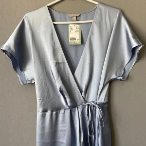Satin blå midi klänning - Helt ny med prislapp kvar. Midi wrapklänning i glansigt gråblått tyg som liknar satin (polyester). Midjemåttet går att justera på insidan med knapp (se bild). Minsta bredd 35 cm. Längd 127 cm. 