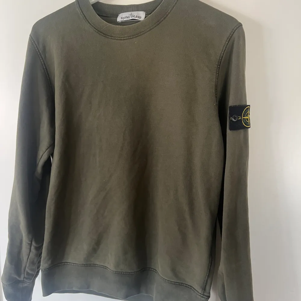 En snygg Stone Island tröja i en mörkgrö/ljus färg. Tröjan har en rund halsringning och en logotyp på vänster arm. storlek M. Hoodies.