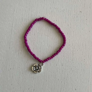 Egengjort pärlarmband  - Ett egengjort pärlarmband med färgeren magenta. Armbandet har en berlock i silver som avbildar en ros. 