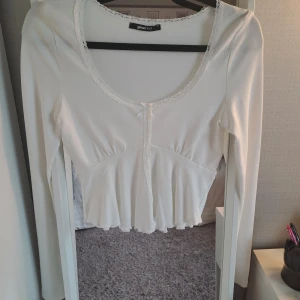 Vit långärmad topp från Gina tricot - En vit långärmad topp från Gina tricot med spetsdetaljer runt halsringningen och en rosett framtill. Toppen har en lätt utsvängd nederkant som ger en feminin touch. Använd en gång, i nyskick🎀✨️