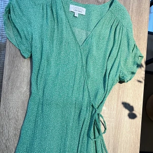 Grön klänning från & Other Stories - Somrig klänning från & Other Stories med små vita prickar. Klänningen har korta ärmar och en omlott-design med knytband i midjan.