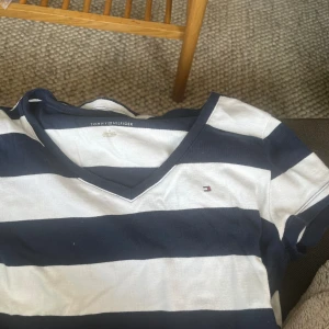 Randig t-shirt från Tommy Hilfiger - En randig t-shirt från Tommy Hilfiger med vita och mörkblå ränder. T-shirten har en v-ringad hals och korta ärmar. Den är tillverkad i ett bekvämt material och har en liten Tommy Hilfiger-logga på bröstet.