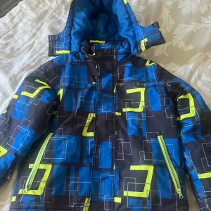 Blå och svart jacka med geometriskt mönster - En blå och svart jacka med geometriskt mönster i grönt och blått. Jackan har en dragkedja framtill och två dragkedjefickor. Den har en huva och är fodrad för extra värme. Ärmarna är lite slitna och ena har två hål som man lätt kan sy ihop. Har en extra knäppning på insidan för att hindra kalluft underifrån.