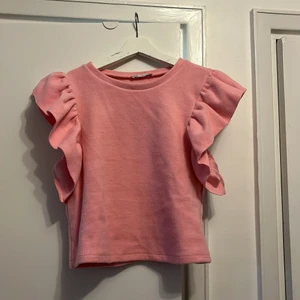 Stickad väst med volanger - Bubbelgumsrosa stickad väst med volanger vid axlarna. Lite kort midja, passar till höga byxor. 