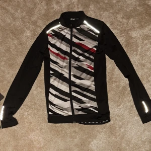 Tränigs jacka/kofta - En andandes tränings jacka med rött, grått och svart mönster. Reflexer vid axlar och på baksidan. I bra skick. Köpare står för frakt.