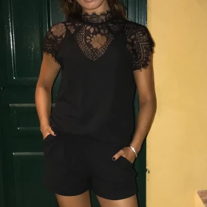 Svart jumpsuit från Neo Noir - En svart jumpsuit från Neo Noir med shorts nertill och fina detaljer i spets upptill. Två fickor på shortsen, som man kan se på första bilden. Knäpps bak i nacken så att den håller sig på plats;) använd vid speciella tillfällen endast därav mkt bra skick!