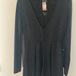 Festig kort byxdress - Svart festig byxdress, nästan lite skimrande i materialet. Djup ringning fram och liten öppning i ryggen. Varsamt använda. Köparen betalar frakt