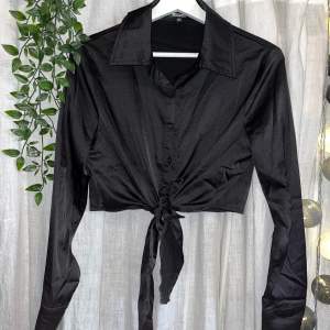 Croppad svart satinskjorta med knappar och knytdetalj framtill. Långa ärmar. Helt i nyskick. Passar både XS och S.