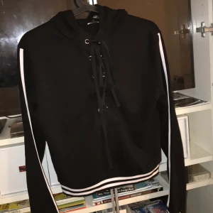 Hoddie kort - Svart kort Hoddie med knytning fram och vita detaljer. Storlek S och inga fickor. 