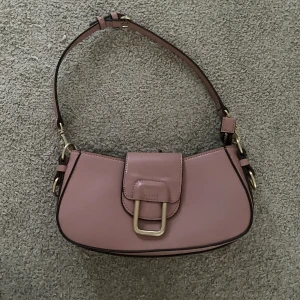 Väska baguette  - Nyskick. Finns två olika band. Ett tjockare i brunt och detta tunna rosa som är på bilden. Fin modell. Är även dragkedja för att stänga.