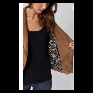 Jacka - Elegant jacka med omlottkrage i mjuk getmocka. Jackan är längre fram och har en tunn quiltad axeldel. Antik-silverdetalj fram, fickor och mudd och ett snyggt leopardmönstrat foder. Färg Almond Nytt pris 3249 kr 