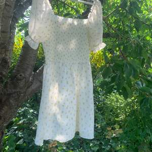 Söt sommar-klänning med smock och liten puffärm som går ner till mitten av låret ungefär. Den är vit med gula blommor och har storlek 44. 