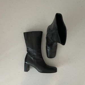 Vintage svarta läderboots med klack - Vintage svarta läderboots med klack. Dragkedja på sidan och i trendig ”squared toe” silhuett! Klacken är 6-7cm hög och den totala höjden på skon är 31cm. Bootsens skostorlek uppskattas vara 39.