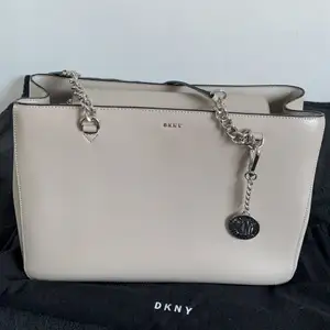 Väska från DKNY i beige/gråaktig färg. Finns ett stort fack med dragkedja mitten samt 4 fack varav 1 har en dragkedja. Finns även ett spänne där man kan hänga fast sina nycklar. Endast använd 5 gånger så den är i nyskick. Medföljer även en svart dustbag.
