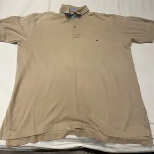 Det är en beige Tommy hilfiger piké tröja som är i storleken L 6/6. Är i bra skick och har inte några märken eller missfärgningar.