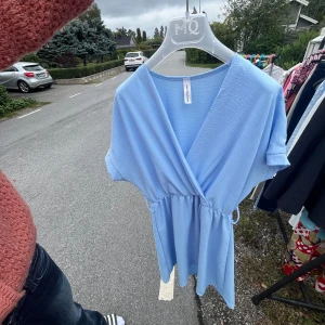 Ljusblå omlottklänning New Collection - Snygg ljusblå kortklänning från New Collection med omlott-design och knyt i midjan. Klänningen har v-ringad hals och korta ärmar, perfekt för varma dagar. Materialet är mjukt och lätt, vilket gör den superbekväm att bära.