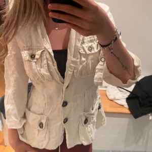 Beige kavaj med fickor och knappar - Snygg beige kavaj med fyra stora fickor framtill och markerad midja. Jackan har stora svarta knappar och trekvartsärmar, perfekt för en chill och stilren look. Materialet känns lätt och passar bra till vår och höst.