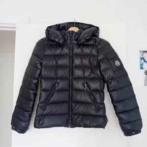 Svart Moncler dunjacka med huva XS - Snygg svart dunjacka från Moncler med huva och dragkedja framtill. Stilren och tidlös design som funkar till allt. Den är i fint begagnat skick, det ända att anmärka är att lappen med info om dun (den med ankorna) börjat lossna, men det är inget som syns när den är på. Inga hål eller fläckar. Finns fler bilder om man vill ha.