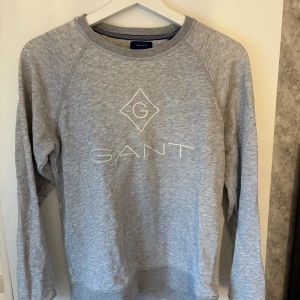 Grå sweatshirt från GANT - Snygg grå sweatshirt från GANT med broderad logga framtill. Tröjan har rund halsringning och långa ärmar. Tillverkad i mjuk bomullsmix som är skön att ha på sig. Perfekt för en chill och stilren look.