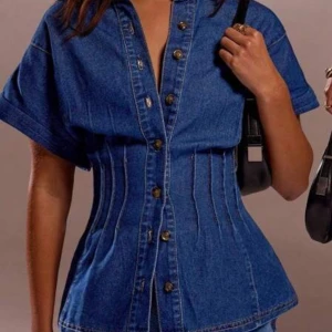 Jeansskjorta med markerad midja - Snygg jeansskjorta i blå denim med korta ärmar och markerad midja för en cool siluett. Skjortan har knappar framtill och dekorativa sömmar som ger en unik look. Perfekt för dig som vill sticka ut med en trendig och modern stil.