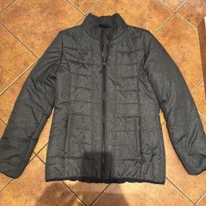 Mörkgrå pufferjacka med dragkedja - Hummel - Säljer en stilren mörkgrå Hummel pufferjacka med hög krage och hel dragkedja framtill. Jackan har två sidofickor och quiltad design som ger en schysst look. Perfekt för kyliga dagar när du vill hålla värmen och ändå se snygg ut. Stolek M