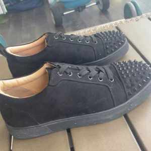 Svarta mockasneakers med nitar Louboutin - Svarta sneakers i mocka från Christian Louboutin med coola svarta nitar över tån. Skorna har snörning, rund tå och svart sula. Insidan är fodrad i beige läder för extra komfort. Perfekta för dig som vill sticka ut med en edgy vibe. Och kommer med ett kort 