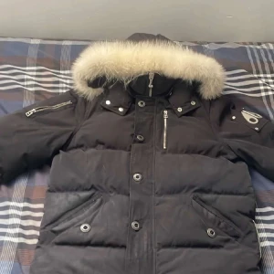 Svart pufferjacka Moose Knuckles - Säljer en svart pufferjacka från Moose Knuckles med fet pälskant på huvan och coola metalldetaljer på ärmen. Jackan har dragkedjor, knappar och flera fickor. Perfekt för kalla dagar och har en riktigt schysst streetstil.