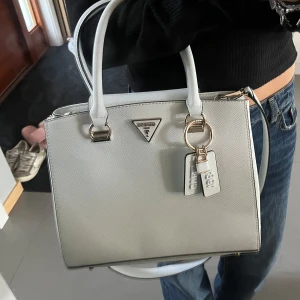Vit Guess väska med najs detaljer - Säljer nu min snygga och stilrena vit handväska från Guess med guldfärgade metalldetaljer och Guess-logga framtill. Väskan har två handtag, avtagbar axelrem, JÄTTEmånga fack vilket typ är det bästa och tre feta charms. Inte en enda defekt.